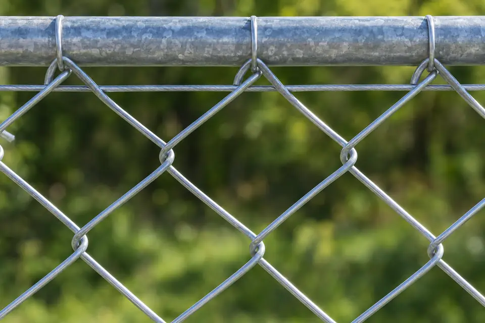 A close-up of galvanized chain link mesh with a top rail and tension wire in sunlight